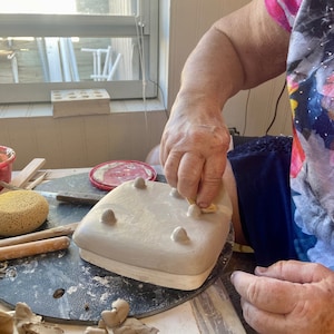 May include: A person is adding small clay pieces to a ceramic dish with five raised bumps. The dish is on a black turntable. There are pieces of clay and tools on the table.
