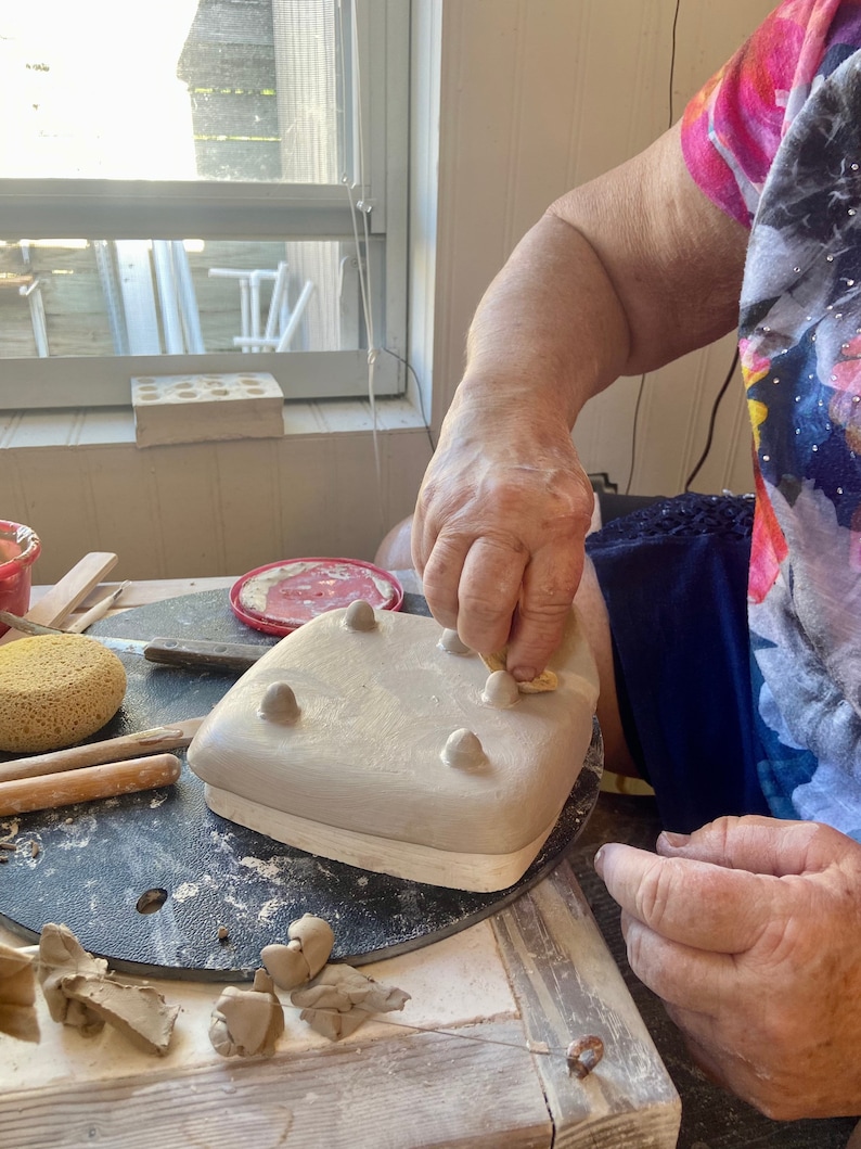 May include: A person is adding small clay pieces to a ceramic dish with five raised bumps. The dish is on a black turntable. There are pieces of clay and tools on the table.