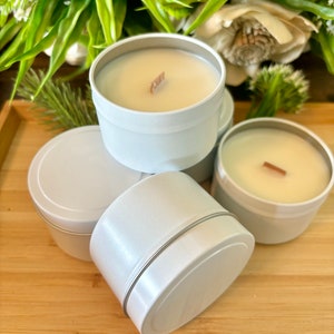 May include: Four white metal candle tins with wooden wicks and a beige wax. The candles are arranged in a pyramid shape on a wooden surface.