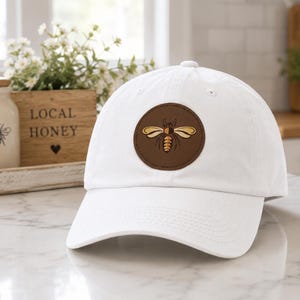 May include: A white baseball cap featuring a circular brown patch on the front. The patch displays a stylized graphic of a bee with yellow and brown wings and a striped body. The cap has a curved brim and a structured crown with eyelet vents.