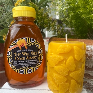 以下が含まれることがあります： 黄色いキャップが付いた蜂蜜のボトルと「Thy Will Bee Done Apiary」と書かれたラベル。その隣には、蝶の模様が入った黄色の蜜蝋キャンドルがあります。蜂蜜のボトルには454gの蜂蜜が入っています。