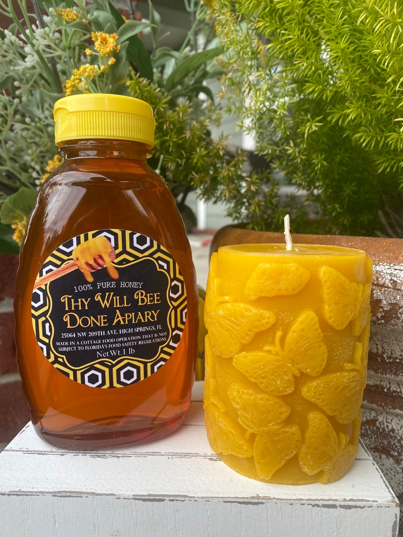 以下が含まれることがあります： 黄色いキャップが付いた蜂蜜のボトルと「Thy Will Bee Done Apiary」と書かれたラベル。その隣には、蝶の模様が入った黄色の蜜蝋キャンドルがあります。蜂蜜のボトルには454gの蜂蜜が入っています。