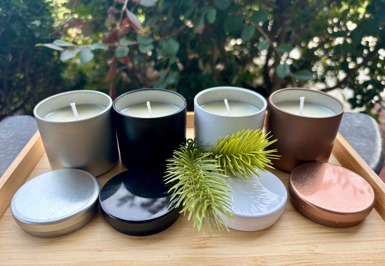 May include: Four cylindrical candles in silver, black, white, and copper metal tins with matching lids. Each candle features a single white wick centered in off-white wax. A small green pine branch rests in front of the white candle container.