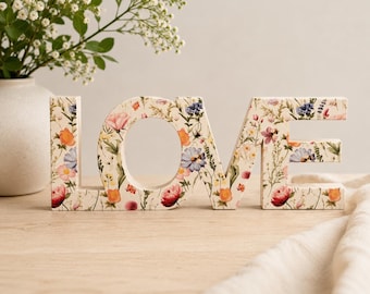 Op de afbeelding: Een vrijstaand houten bord dat het woord LOVE in hoofdletters spelt. Het oppervlak van de letters is voorzien van een kleurrijke botanische print met diverse wilde bloemen, waaronder roze, oranje en blauwe bloesems op een crèmekleurige achtergrond.