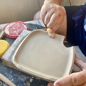 May include: A person is smoothing the edges of a ceramic plate with a damp sponge. The plate is light gray and has a slightly rounded edge. The plate is sitting on a wooden surface.