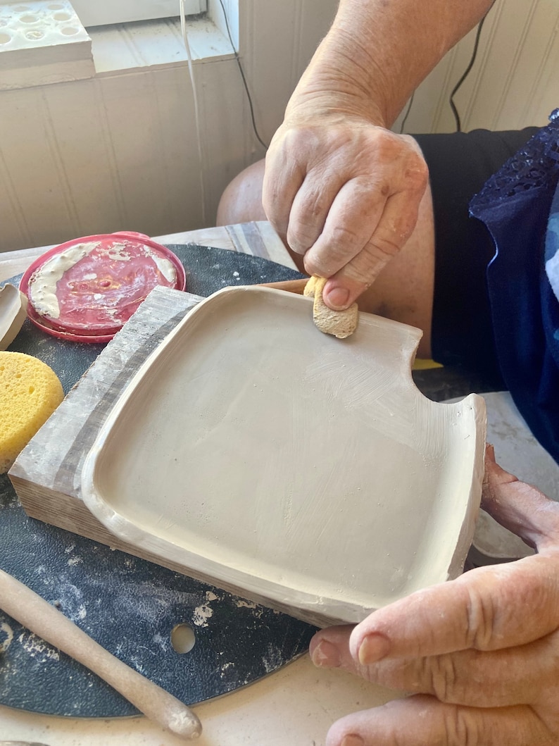 May include: A person is smoothing the edges of a ceramic plate with a damp sponge. The plate is light gray and has a slightly rounded edge. The plate is sitting on a wooden surface.
