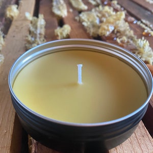 May include: A round, metal tin candle with a pale yellow wax and a white wick. The candle sits on a wooden surface, with pieces of honeycomb in the background. The candle is a natural product.