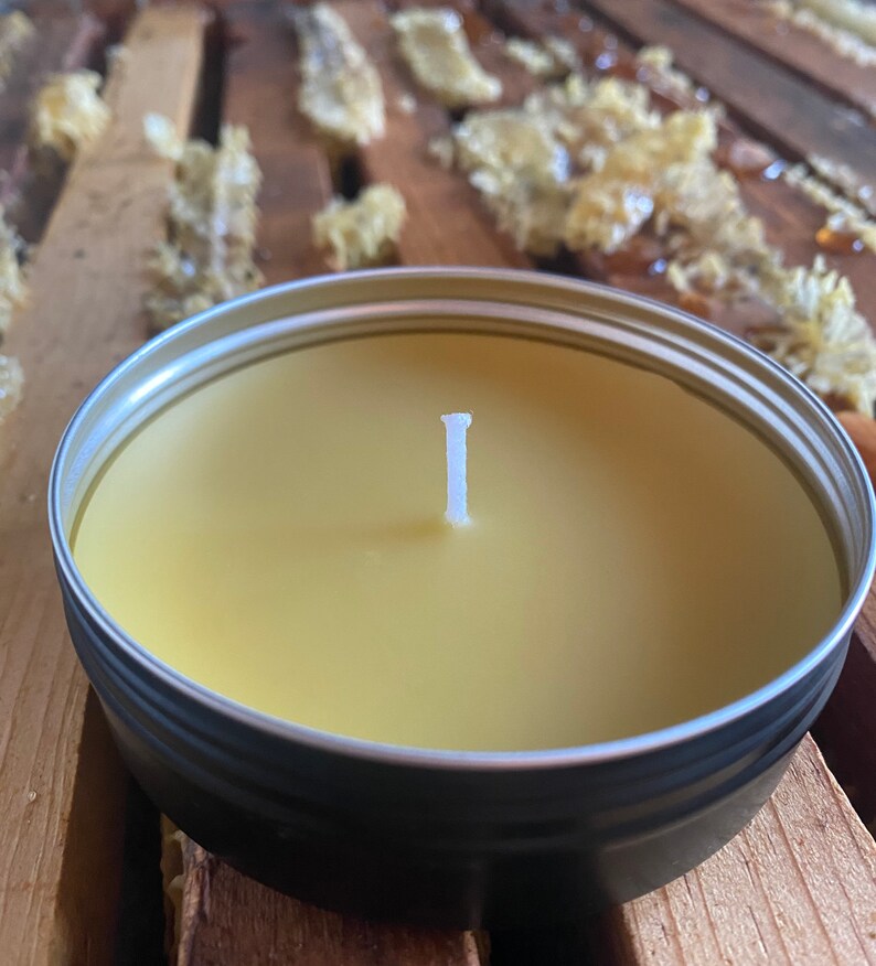 May include: A round, metal tin candle with a pale yellow wax and a white wick. The candle sits on a wooden surface, with pieces of honeycomb in the background. The candle is a natural product.