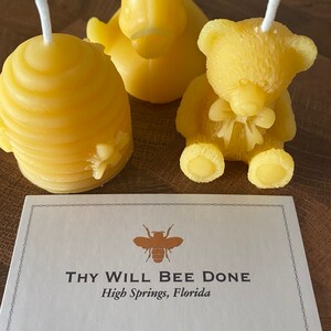 May include: Three yellow beeswax candles in the shapes of a beehive, a duck, and a teddy bear. The teddy bear has a bow. A card with the text "THY WILL BEE DONE" and a bee graphic is in the foreground.