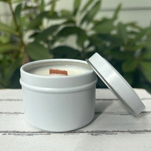 May include: A white metal candle tin with a matching lid resting beside it. The container is filled with smooth white wax and features a single rectangular wooden wick centered in the middle, providing a clean and simple aesthetic for the candle.
