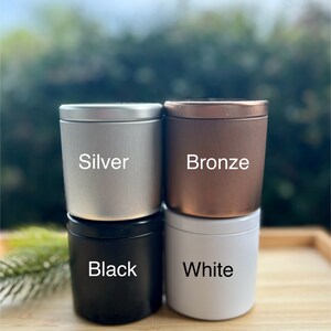 May include: Four cylindrical metal tins with lids, arranged in a two-by-two stack. The top left tin is silver, the top right is bronze, the bottom left is black, and the bottom right is white. Each tin features its color name printed in white text.