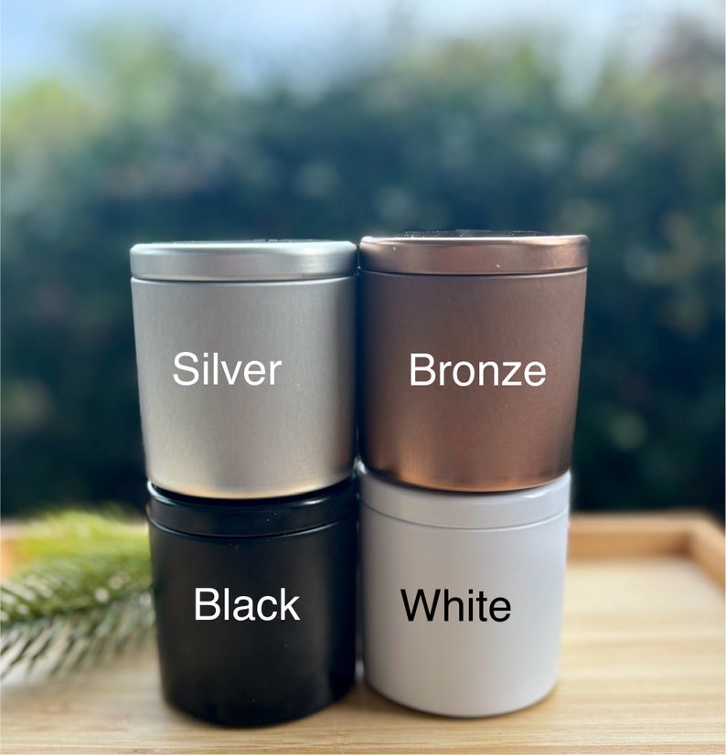 May include: Four cylindrical metal tins with lids, arranged in a two-by-two stack. The top left tin is silver, the top right is bronze, the bottom left is black, and the bottom right is white. Each tin features its color name printed in white text.