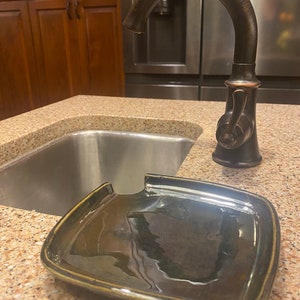 May include: A black ceramic dish with a cut-out for a faucet spout. The dish is sitting on a brown countertop next to a stainless steel sink.