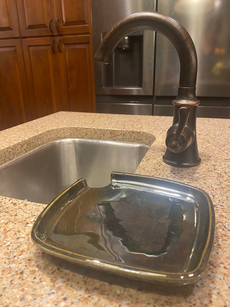 May include: A black ceramic dish with a cut-out for a faucet spout. The dish is sitting on a brown countertop next to a stainless steel sink.