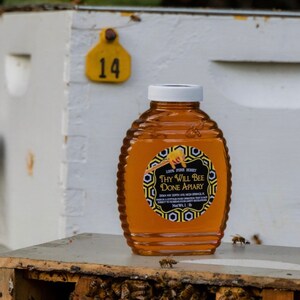 May include: A bottle of honey from Thy Will Bee Done Apiary, with a white cap and a label. The label reads "100% Pure Honey". The bottle is sitting on a wooden surface with bees. A yellow tag with the number 14 is visible.
