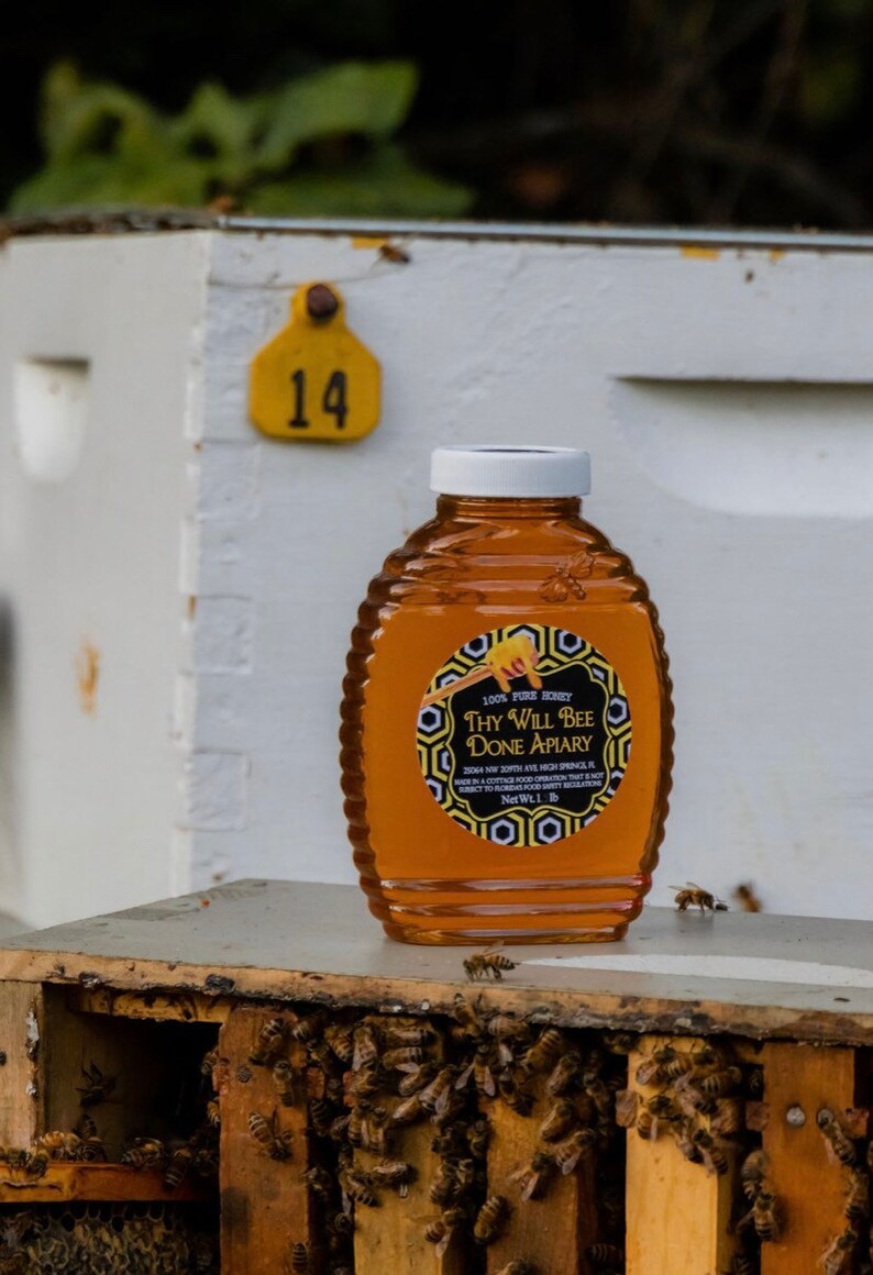 May include: A bottle of honey from Thy Will Bee Done Apiary, with a white cap and a label. The label reads "100% Pure Honey". The bottle is sitting on a wooden surface with bees. A yellow tag with the number 14 is visible.