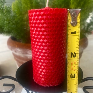 May include: A cylindrical red beeswax candle with a textured honeycomb pattern and a single white wick. A yellow measuring tape is positioned vertically against the side of the candle, showing a height of approximately four inches.