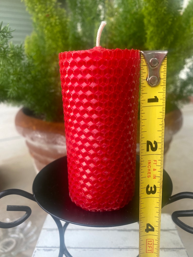 May include: A cylindrical red beeswax candle with a textured honeycomb pattern and a single white wick. A yellow measuring tape is positioned vertically against the side of the candle, showing a height of approximately four inches.