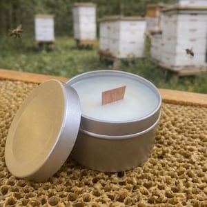 May include: A round silver metal candle tin with a matching lid placed to the side. The candle contains white wax and a single rectangular wooden wick centered in the middle. The container is resting on a textured, golden-brown honeycomb surface.