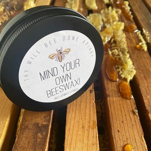 May include: A black tin of beeswax with a white label that reads "MIND YOUR OWN BEESWAX!" and "THY WILL BEE DONE APIARY". The tin sits on wooden slats with honeycomb and honey droplets. The product is from High Springs, Florida.