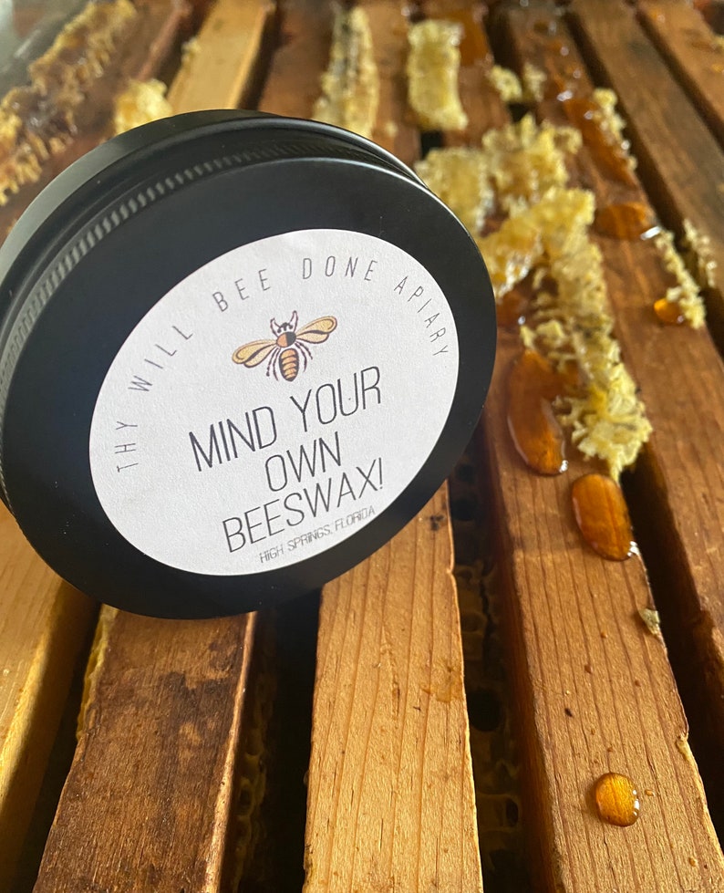 May include: A black tin of beeswax with a white label that reads "MIND YOUR OWN BEESWAX!" and "THY WILL BEE DONE APIARY". The tin sits on wooden slats with honeycomb and honey droplets. The product is from High Springs, Florida.