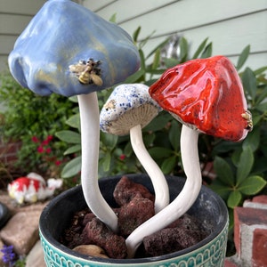 May include: Three ceramic mushrooms in a blue and green planter. The mushrooms are painted in blue, red, and white with black speckles. The planter has a floral design.