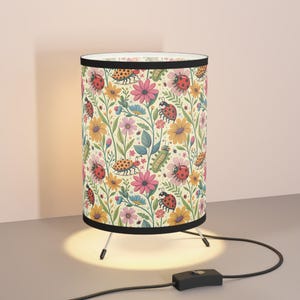 May include: A cylindrical table lamp with a black trim and tripod base. The lamp shade features a repeating pattern of red ladybugs, yellow and pink flowers, and green leaves on a cream background. The lamp is plugged into a wall outlet.