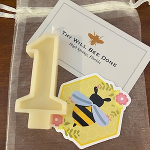May include: A cream-colored number one candle with a flat base, a sticker with a bee graphic, and a card that reads "THY WILL BEE DONE" in a sheer drawstring bag. The bee sticker is on a yellow honeycomb background.