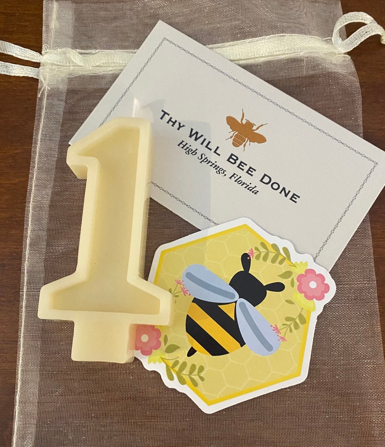 May include: A cream-colored number one candle with a flat base, a sticker with a bee graphic, and a card that reads "THY WILL BEE DONE" in a sheer drawstring bag. The bee sticker is on a yellow honeycomb background.