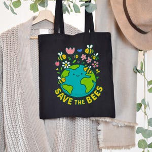 May include: Black tote bag featuring a colorful graphic of the Earth with a smiling face, surrounded by bees and flowers. The text "SAVE THE BEES" is printed in yellow. The bag has black handles and is hanging on a wooden hanger.