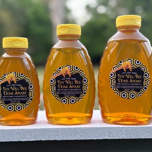 May include: Three glass bottles of honey, each with a yellow lid and a black and gold label that reads "100% Pure Honey Thy Will Bee Done Apiary". The bottles are arranged in a row from smallest to largest.