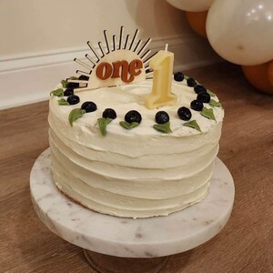 May include: A white frosted cake decorated with blueberries and green leaves, topped with a wooden sunburst design that reads "one" and a number one candle. The cake sits on a marble cake stand.