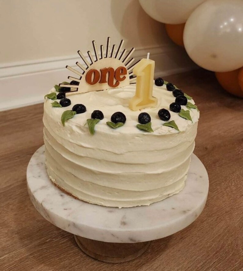 May include: A white frosted cake decorated with blueberries and green leaves, topped with a wooden sunburst design that reads "one" and a number one candle. The cake sits on a marble cake stand.