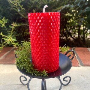 May include: A cylindrical red candle with a textured honeycomb pattern on its surface and a single white wick at the top. The candle sits on a black metal stand with curved legs, accompanied by a small sprig of green foliage at its base.