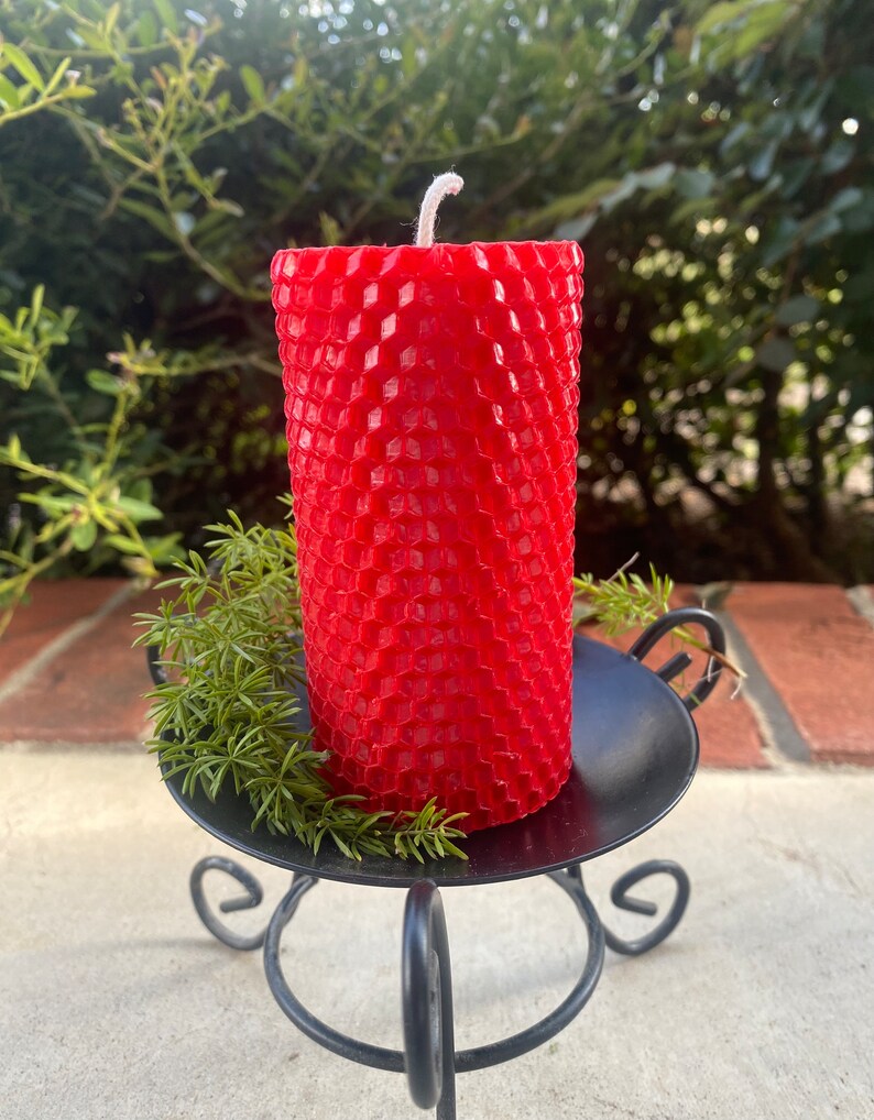 May include: A cylindrical red candle with a textured honeycomb pattern on its surface and a single white wick at the top. The candle sits on a black metal stand with curved legs, accompanied by a small sprig of green foliage at its base.