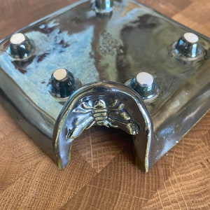 May include: A dark blue ceramic planter with a raised, arched opening and four white ceramic feet. The planter has a decorative design of a stylized insect on the front.