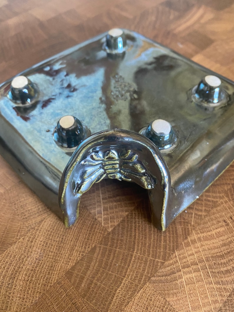 May include: A dark blue ceramic planter with a raised, arched opening and four white ceramic feet. The planter has a decorative design of a stylized insect on the front.