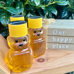 Peut inclure: Trois petits pots de miel en verre en forme d'ours avec des couvercles jaunes. Les pots sont étiquetés "Gallberry" et sont posés sur une surface en bois. Le texte "Our happy place" est visible sur une pancarte en bois derrière les pots.