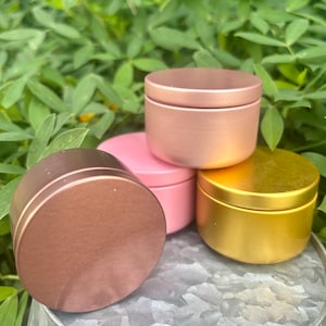 May include: Four round metal tins with lids. The tins are stacked on top of each other. The top tin is rose gold, the second tin is pink, the third tin is gold, and the bottom tin is a darker rose gold.