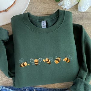 May include: A dark green sweatshirt with a line of five cartoon bees in yellow and black, each with a different expression. The bees are holding flowers and a honeycomb.