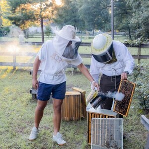 以下が含まれることがあります： 養蜂用の防護服と帽子を着用した2人が、ミツバチの巣箱を扱っています。一人は煙を出す道具を持ち、もう一人はハニカムフレームを検査しています。木製の箱と金網も見えます。