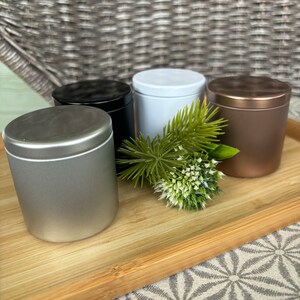 May include: A set of four cylindrical metal tins with lids in silver, black, white, and copper finishes. The containers are arranged together with a small decorative sprig of green pine needles and a white flower head placed in the center.