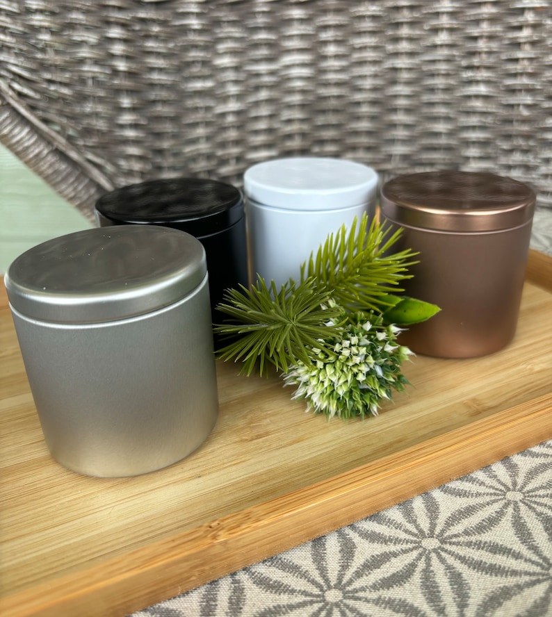 May include: A set of four cylindrical metal tins with lids in silver, black, white, and copper finishes. The containers are arranged together with a small decorative sprig of green pine needles and a white flower head placed in the center.