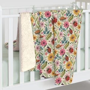 May include: A floral patterned blanket with a cream-colored sherpa backing. The blanket features a repeating pattern of pink, yellow, and blue flowers, green leaves, ladybugs, and caterpillars. The blanket is draped over a white crib.