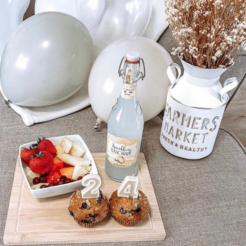 May include: A birthday celebration scene with white balloons, a bottle of sparkling lemonade, and two muffins with candles that read "24." A wooden cutting board holds a bowl of fruit and a white "Farmers Market" vase with dried flowers.
