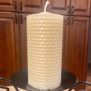 May include: A cylindrical off-white candle with a textured honeycomb pattern on its exterior surface. The candle features a single white cotton wick protruding from the center of the top flat surface and rests on a black metal candle holder.