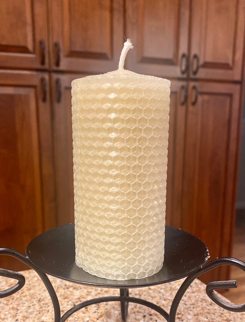 May include: A cylindrical off-white candle with a textured honeycomb pattern on its exterior surface. The candle features a single white cotton wick protruding from the center of the top flat surface and rests on a black metal candle holder.