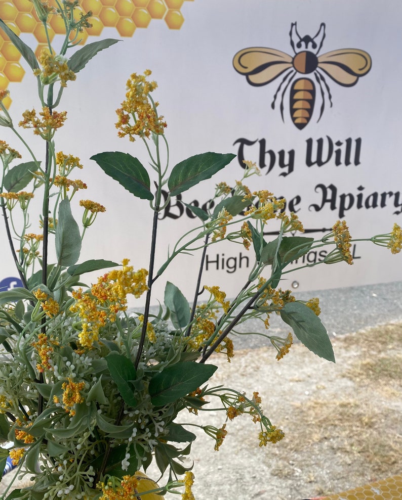May include: A bouquet of artificial yellow and orange flowering stems with green leaves positioned in front of a banner featuring a bee graphic and the text Thy Will Bee Done Apiary High Springs FL with a honeycomb pattern in the corner.