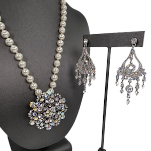 May include: A pearl necklace with a large, ornate pendant featuring iridescent crystals. Matching chandelier earrings with dangling crystals are displayed on a stand. The jewellery set is presented on a dark grey mannequin.