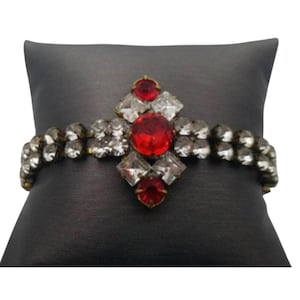 May include: A vintage bracelet featuring a central design of red and clear gemstones. The bracelet has a gold-tone metal setting and is adorned with a large, round red stone at the center, surrounded by clear, square-cut stones. The band is composed of smaller, clear stones.